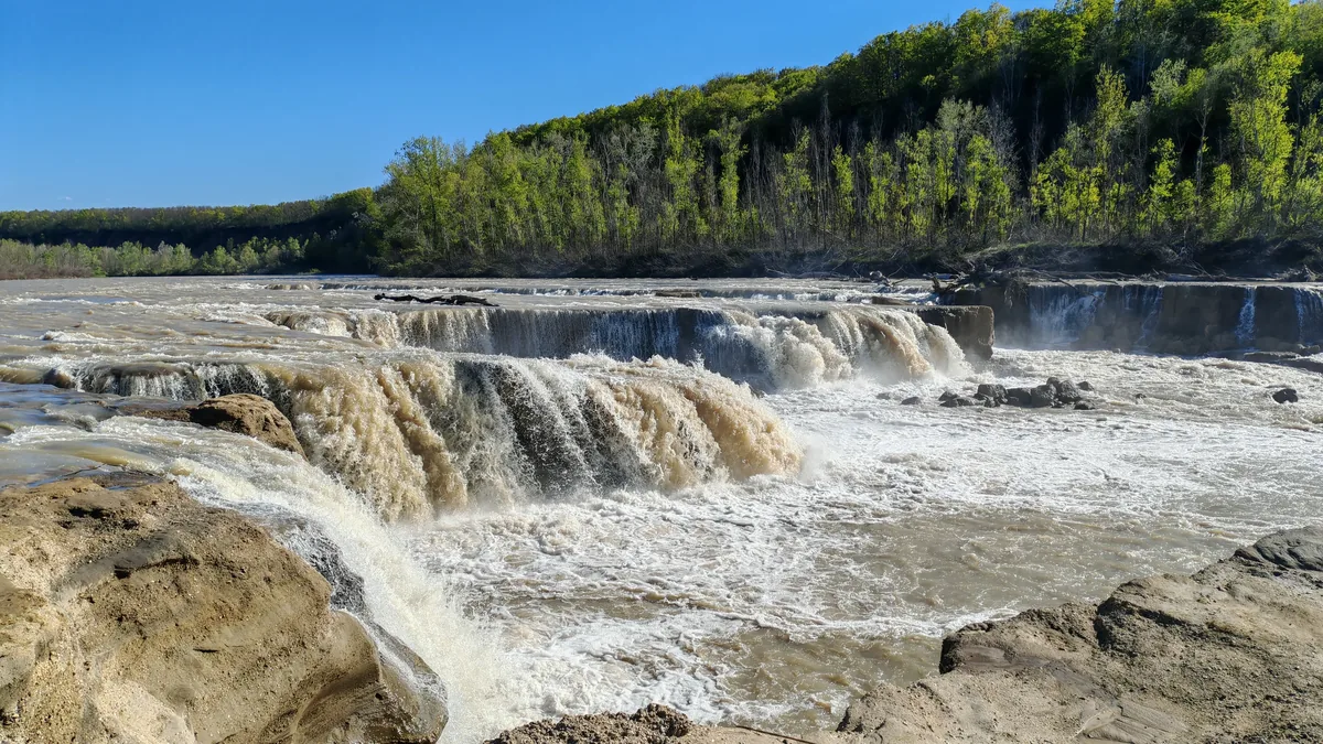 Широкий каскад на реке Белой с каменными уступами и зелёным лесом за водой.